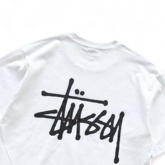White BASIC STÜSSY TEE t-shirt displayed on a flat surface with a simple design and logo.